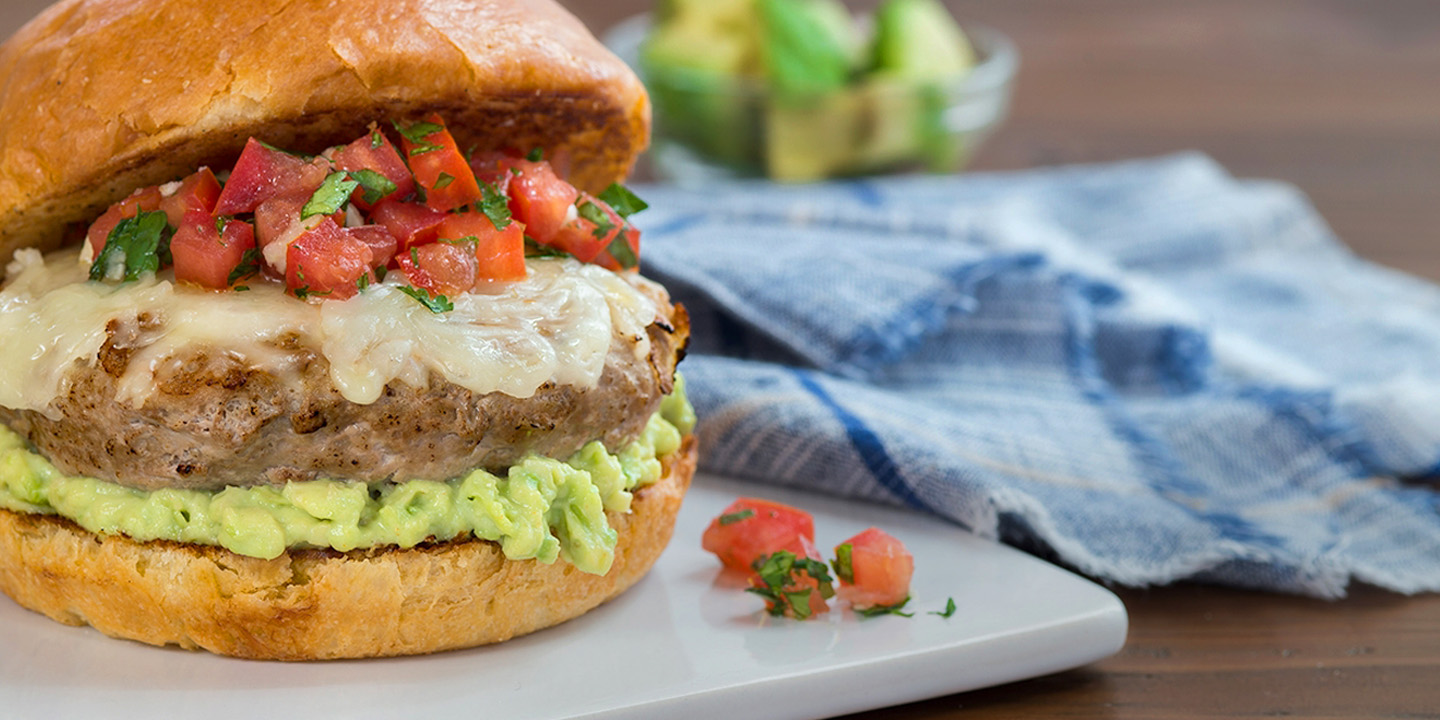 Turkey burger on a plate topped with bruschetta, smashed avocado, and melted cheese